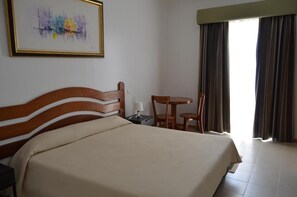 Una habitación de hotel con una cama grande, un cabecero de madera, una mesita con una silla, una mesita de noche y una ventana con cortinas.