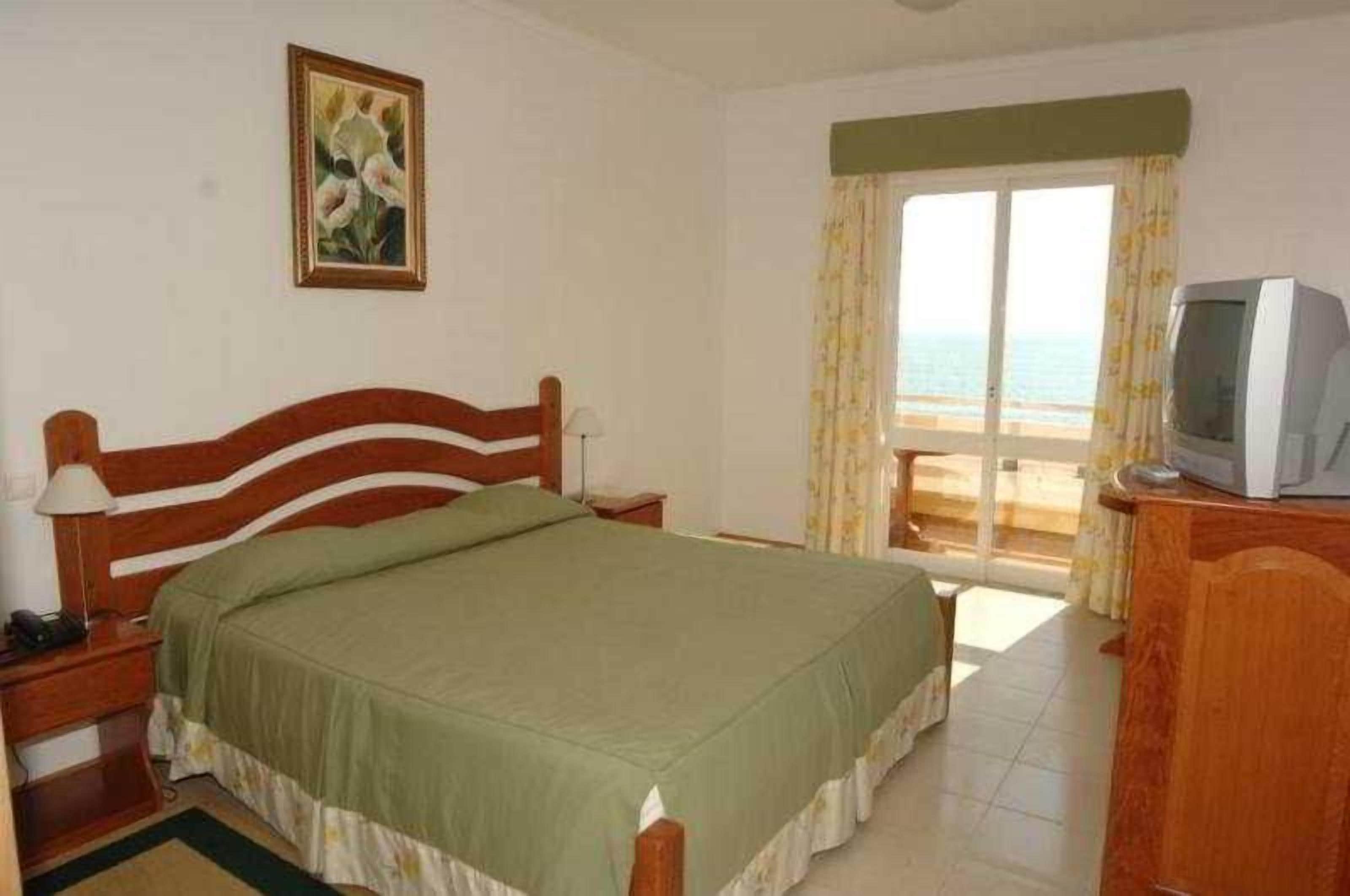 A room with a bed, a television, and a view of the sea through a window.