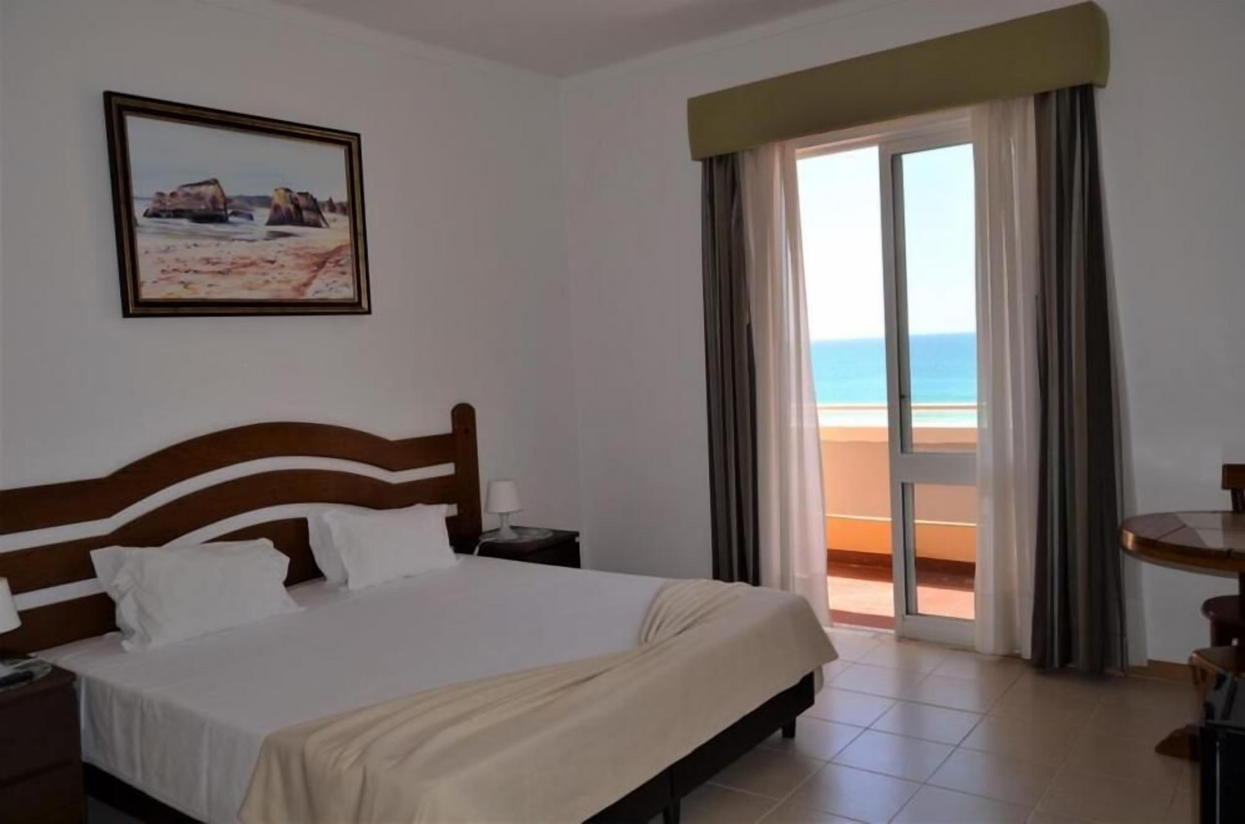 Ein Hotelzimmer mit Bett, Nachttisch, einem Gemälde, einem Fenster mit Vorhängen und Blick auf den Strand.