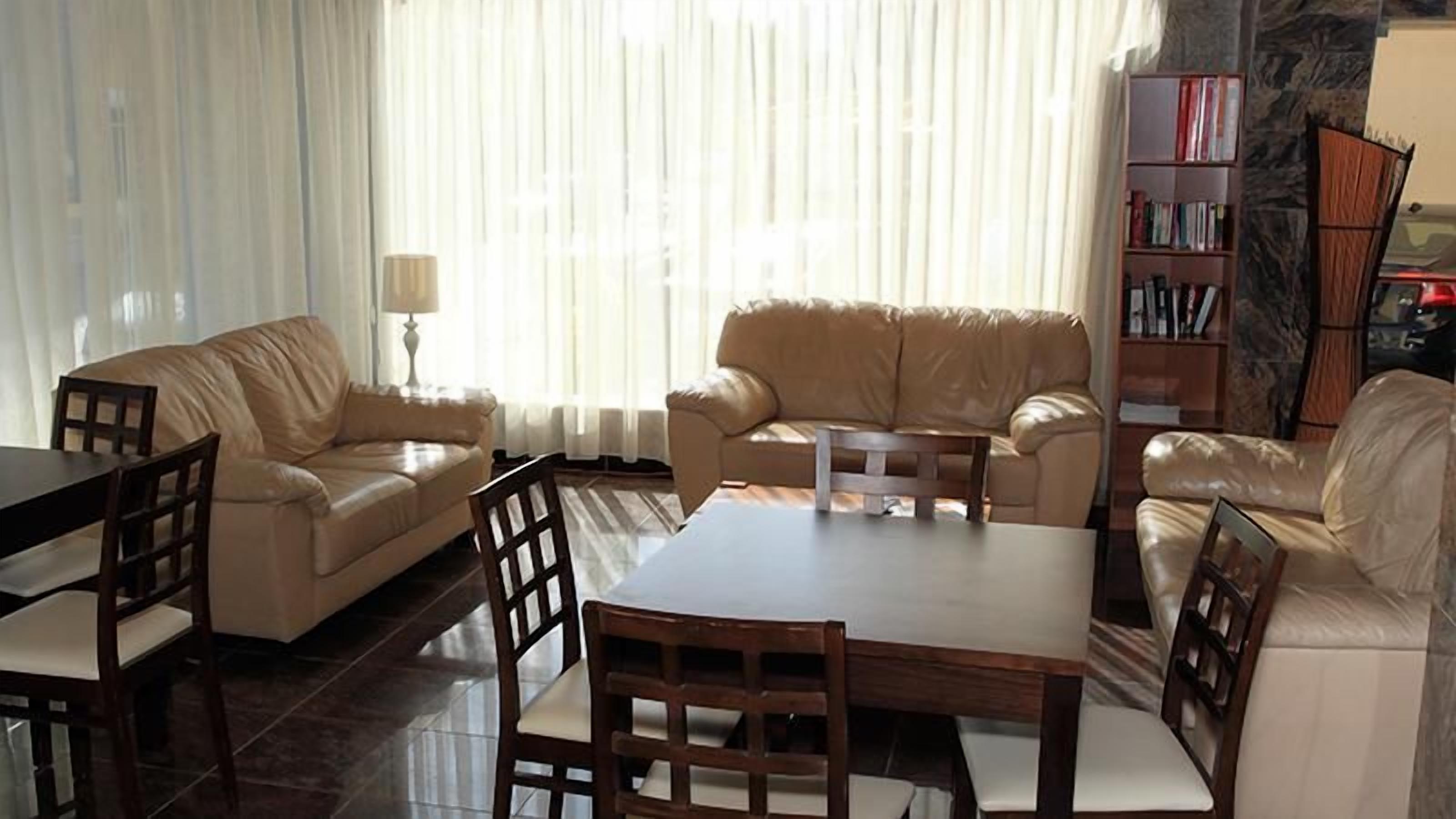 A living room with a sofa, a dining table, and chairs. There is a bookshelf and a lamp in the room.