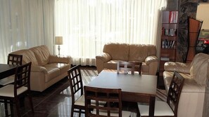 Una sala de estar con un sofá, una mesa de comedor y sillas. Hay una estantería y una lámpara en la habitación.