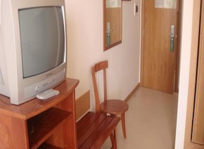 A room with a wooden chair, a television on a wooden stand, and a door with a mirror and a light switch.