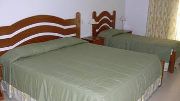 A room with two wooden beds, each with a green bedspread and a floral-patterned skirt. There are bedside tables with lamps and a curtain with a yellow and white pattern.