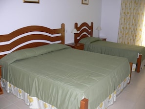 Une chambre avec deux lits en bois, chacun recouvert d’une couverture verte et orné d’une jupe de lit à motifs floraux. On y trouve des tables de chevet avec des lampes, ainsi qu’un rideau à motifs jaunes et blancs.