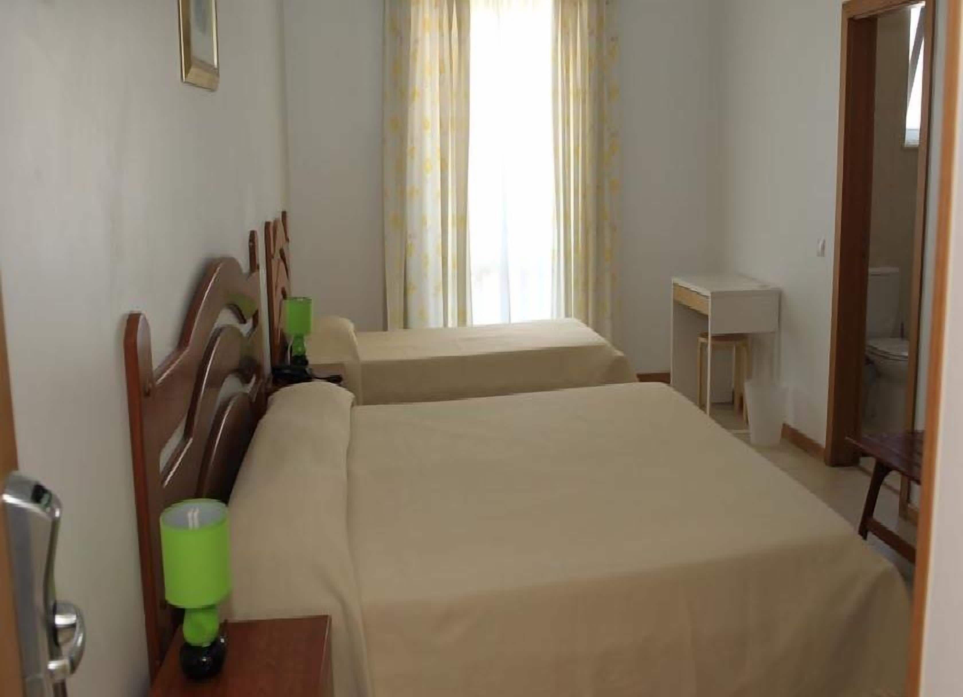 Habitación de hotel con dos camas, cabecero de madera, mesita de noche con una lámpara verde, ventana con cortinas y otra mesita de noche con una lámpara blanca.