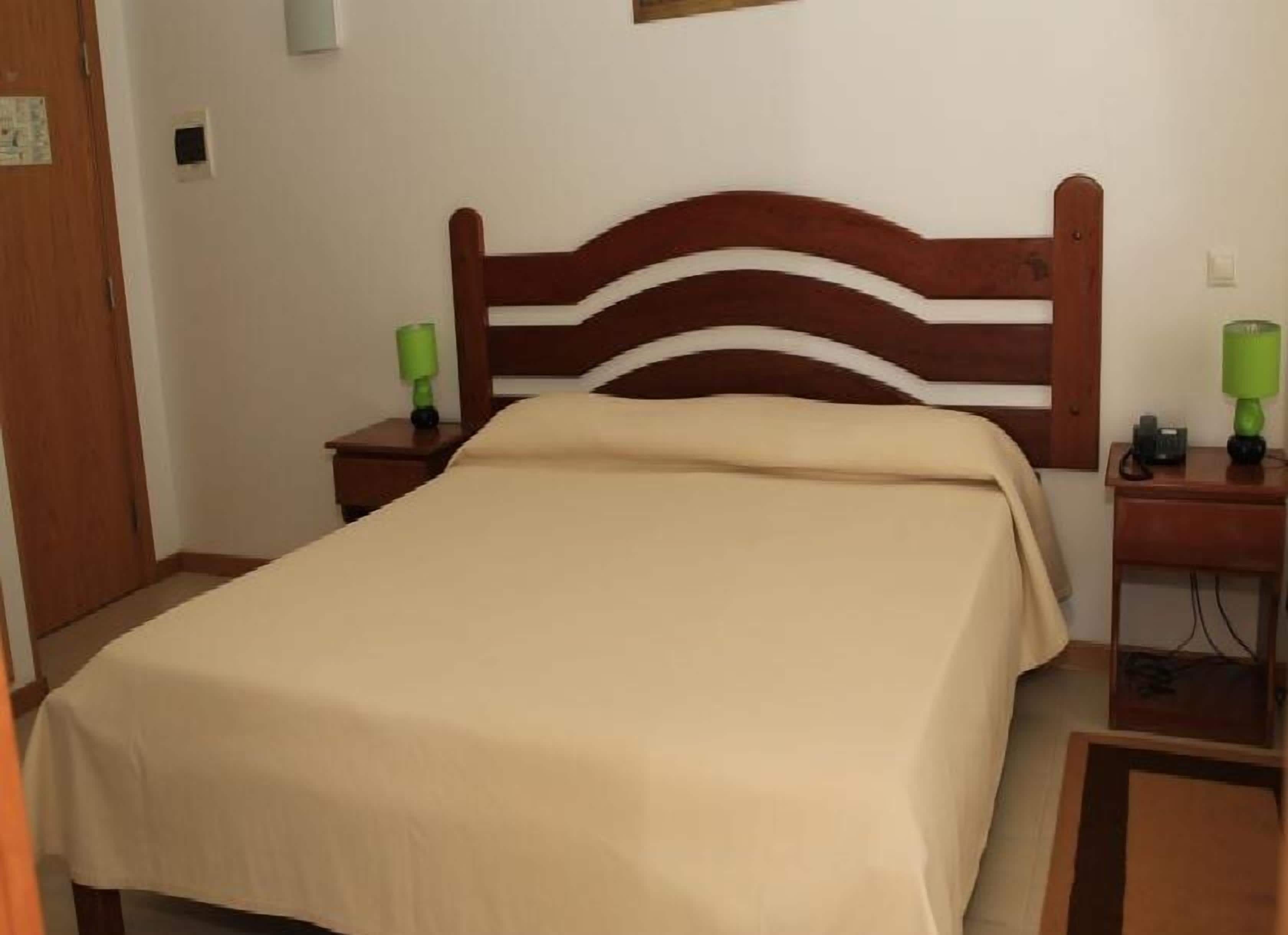 A neatly made bed with a wooden headboard, two bedside tables with lamps, and a plain wall.