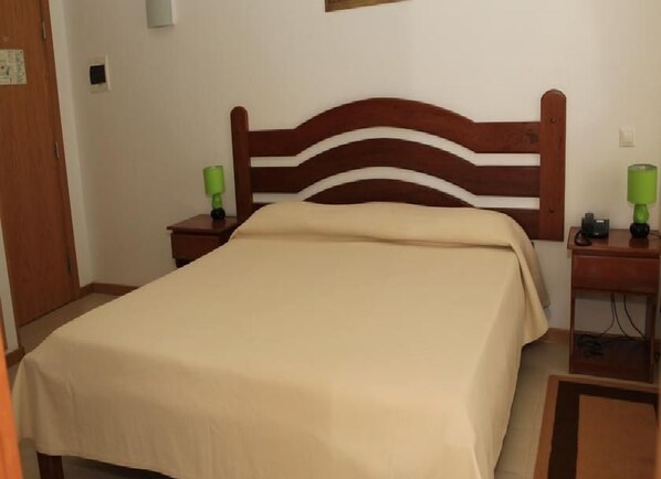 Una cama bien hecha con cabecera de madera, dos mesitas de noche con lámparas y una pared lisa.
