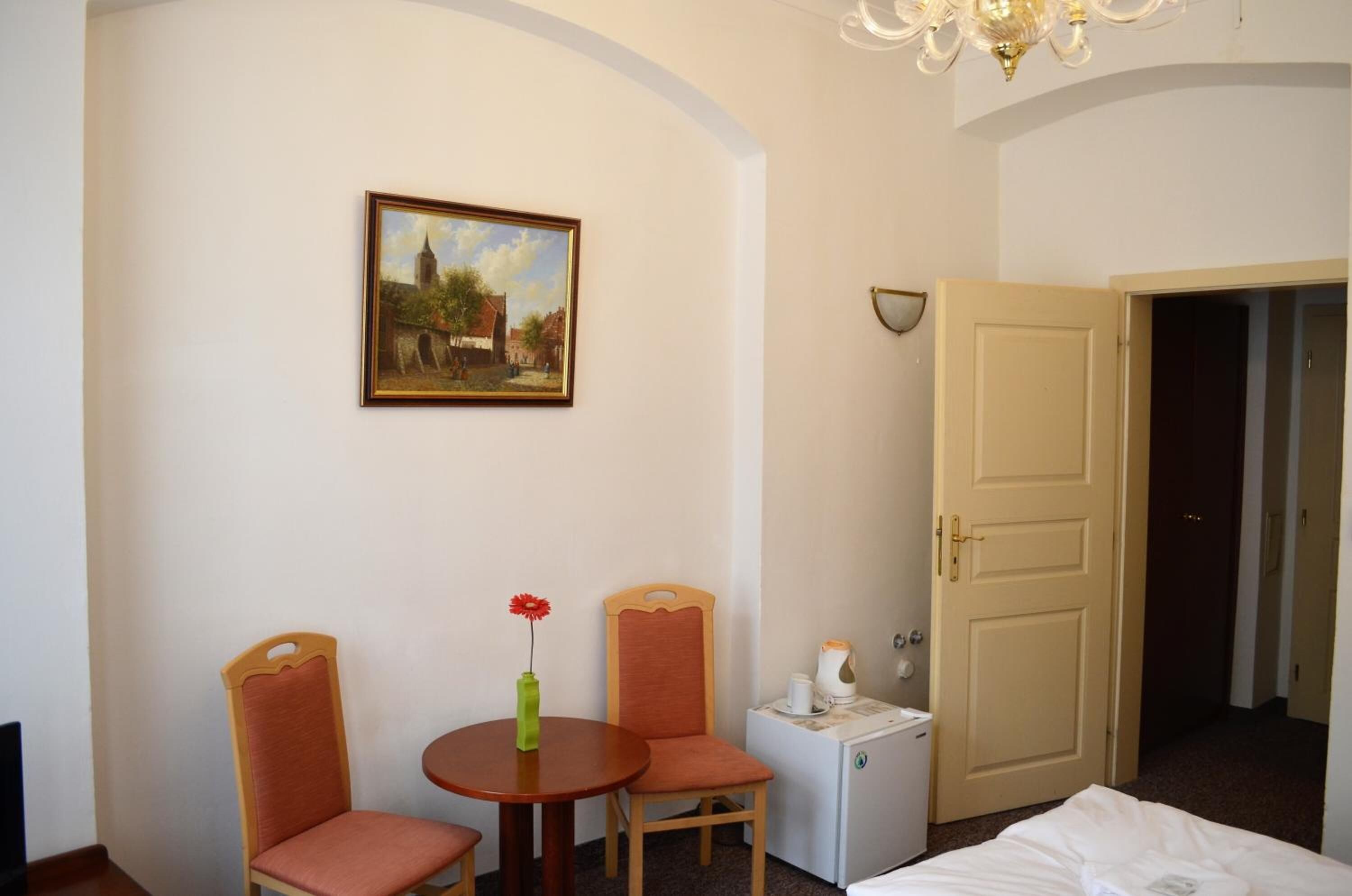 Habitación de hotel con cama, una mesita pequeña con dos sillas, un cuadro en la pared, una puerta y una mesita de noche con lámpara.