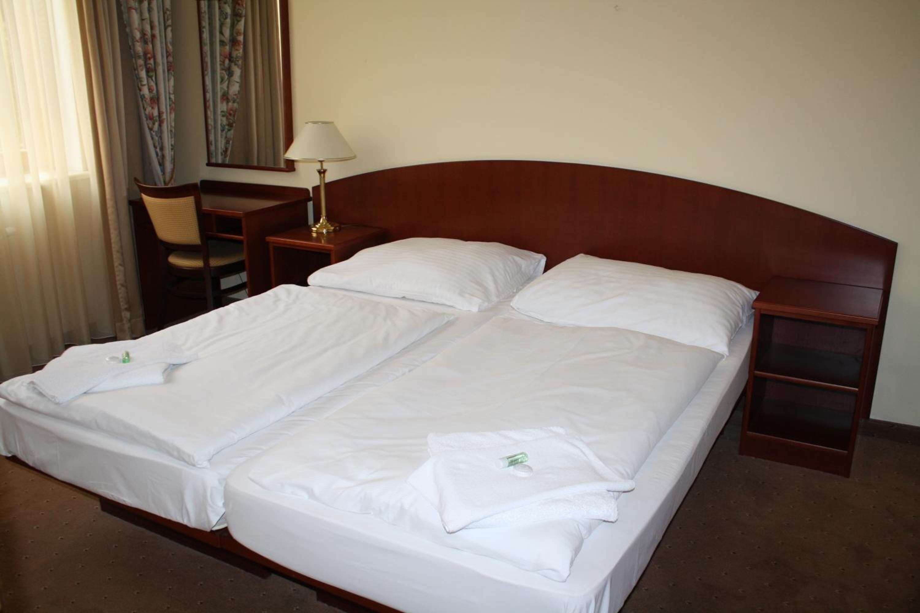 A double bed with white linens, a wooden headboard, a bedside table with a lamp, and a chair.