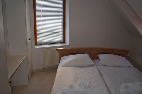 Una cama individual con ropa de cama blanca y dos almohadas, cabecera de madera, ventana con persianas, radiador y armario blanco.