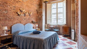 Una habitación con pared de ladrillo, una cama grande con ropa de cama azul, una araña, una ventana con contraventanas abiertas y un piso de baldosas.