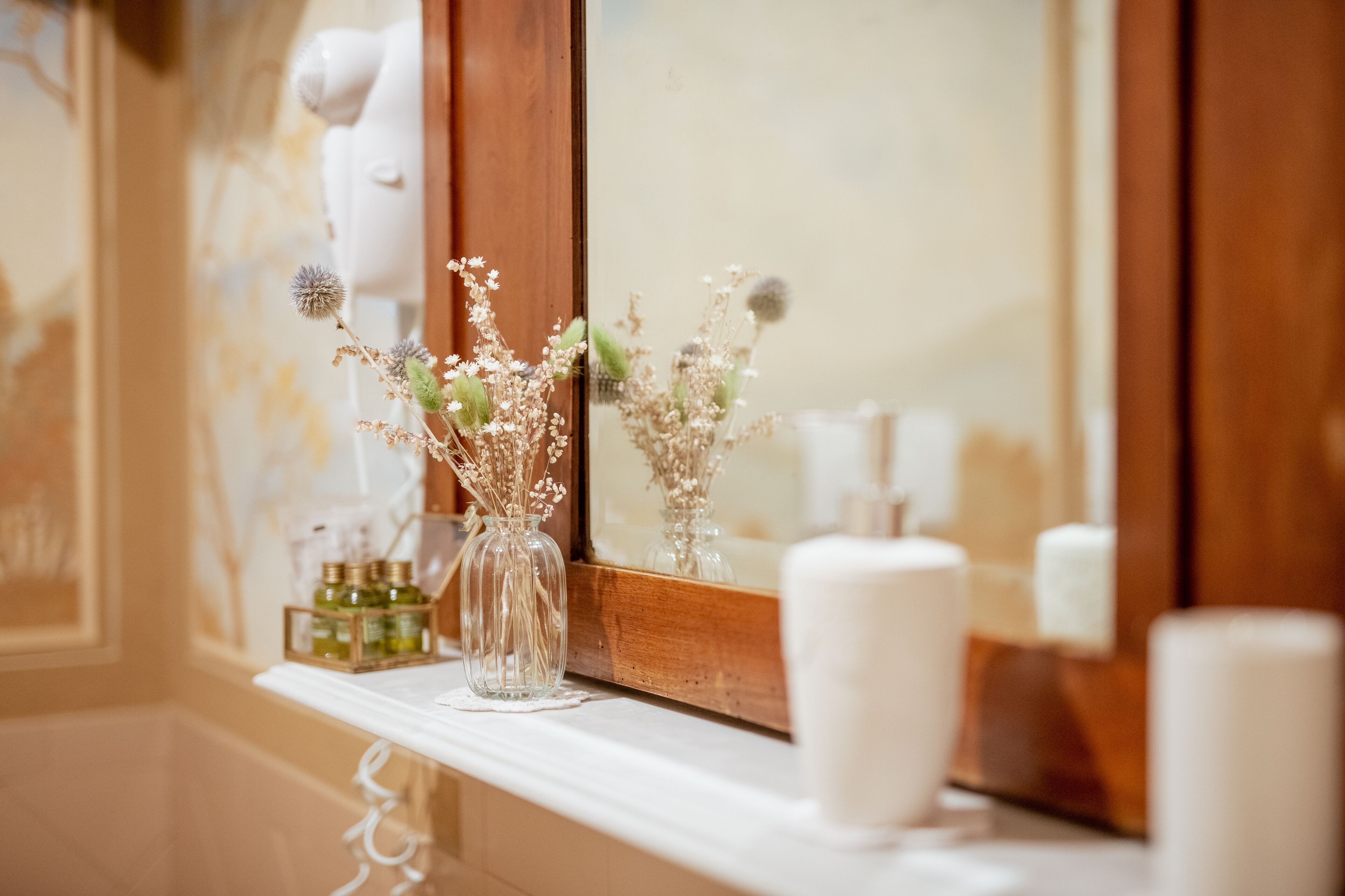 Un tocador de baño con un espejo, un jarrón con flores secas y una botella de loción.
