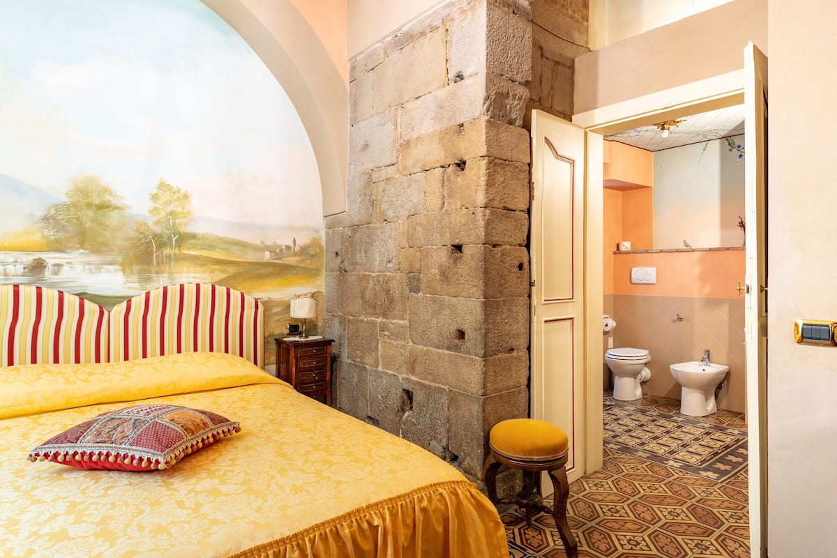 Una stanza con un letto, una parete in pietra, un bagno con il water e un bidet, e un affresco raffigurante un paesaggio.