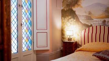 A bedroom with a bed, a nightstand with a lamp, a wall mural, and a door with stained glass windows.