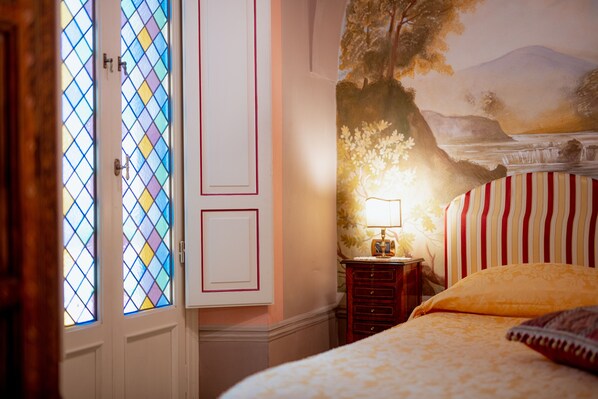 Un dormitorio con cama, mesita de noche con lámpara, un mural en la pared y una puerta con vidrieras.