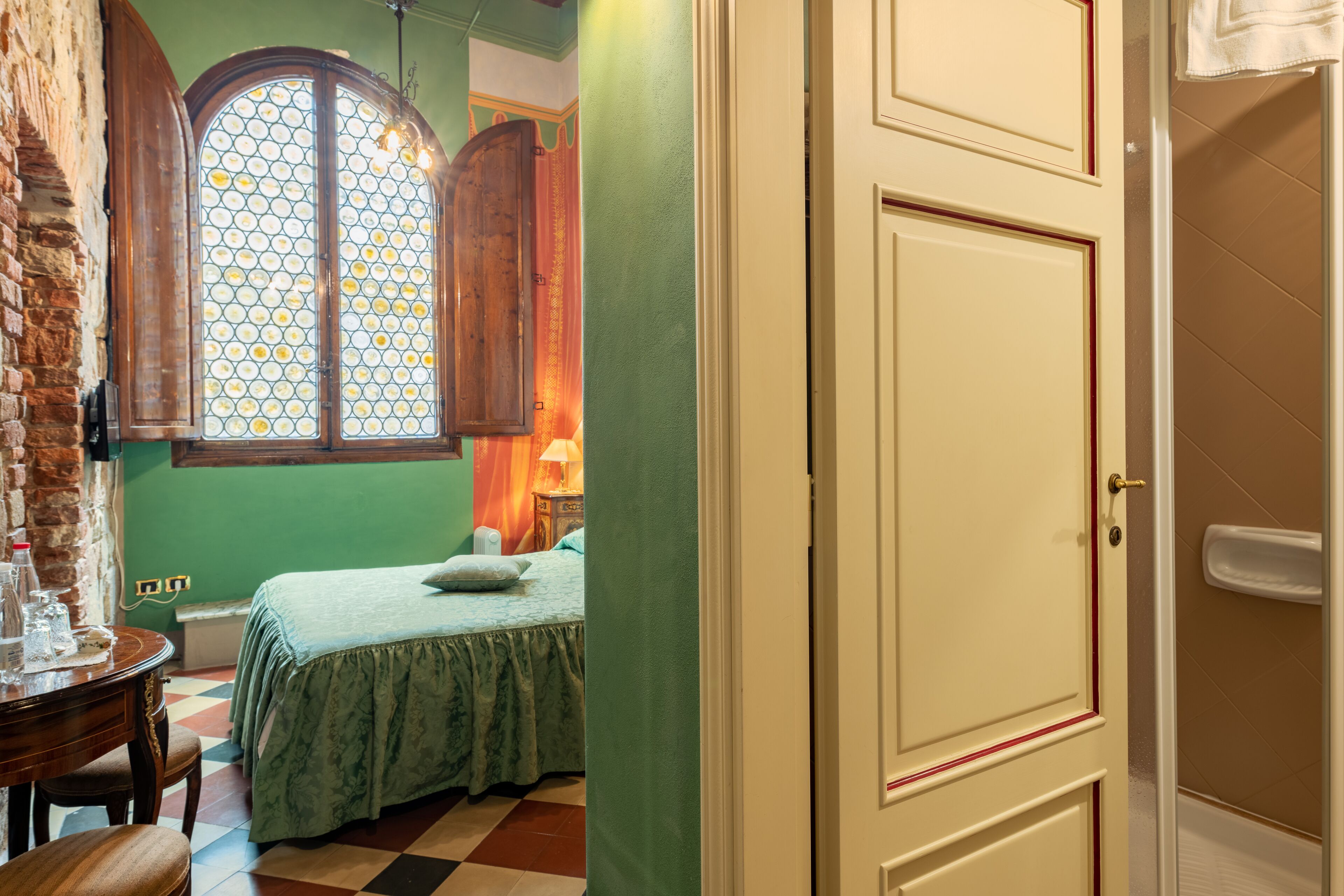 A room with a checkered floor, a bed with a green bedspread, a wooden cabinet, and a window with a diamond pattern.