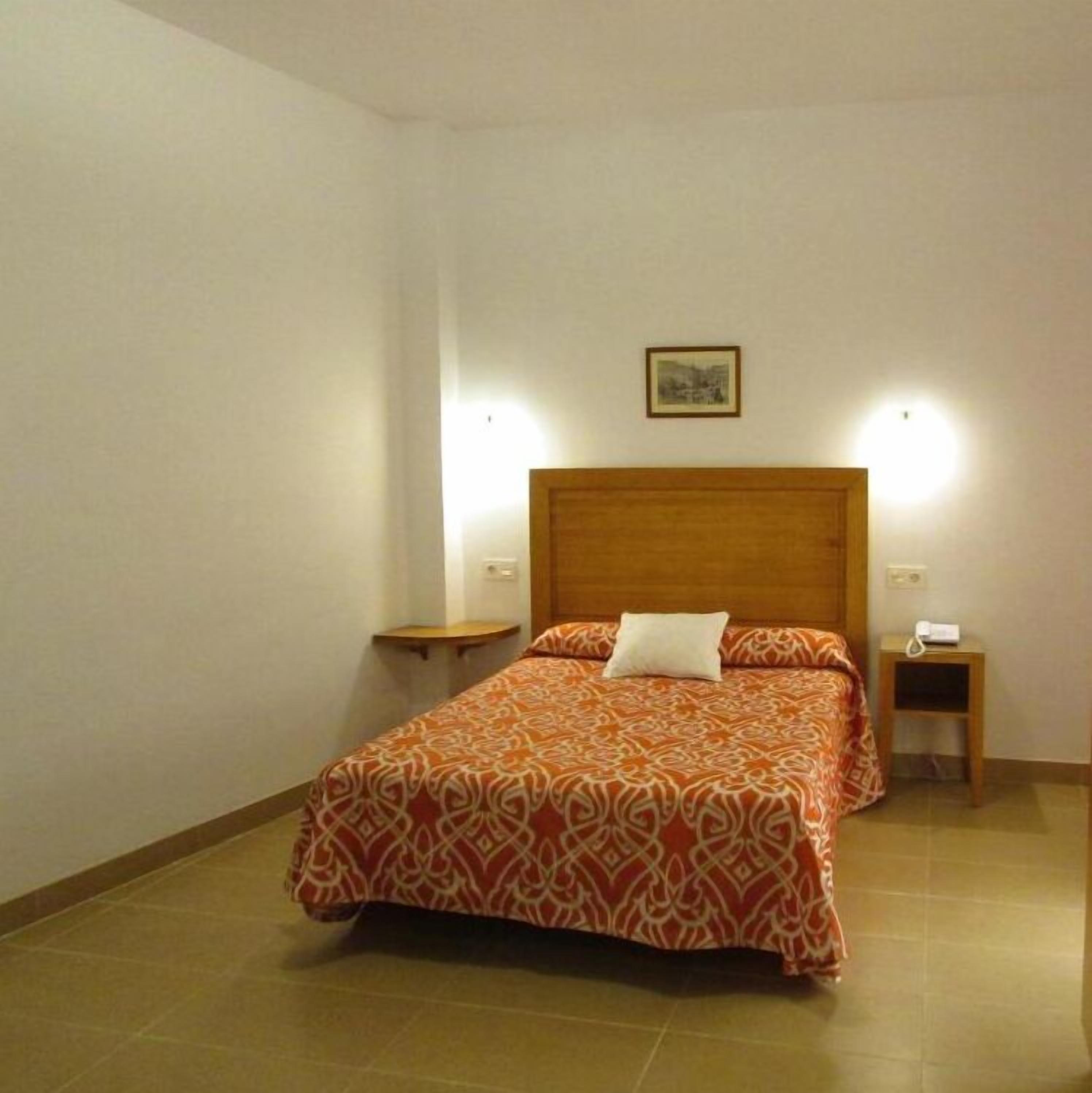 Una cama individual con colcha estampada, dos mesitas de noche, una lámpara de pared y un pequeño cuadro en la pared.