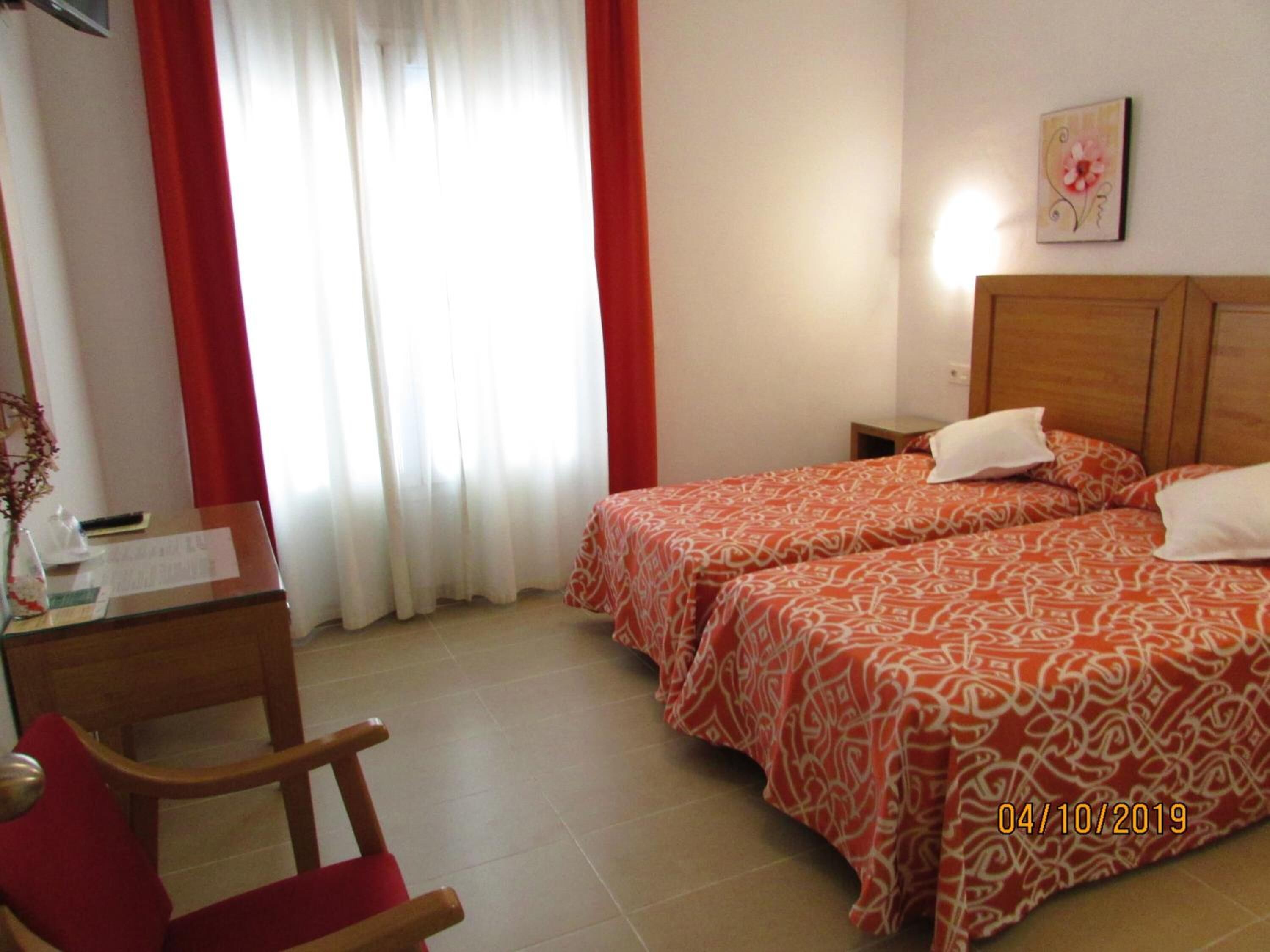 Una camera d'albergo con due letti, una scrivania con sedia, un comodino e una finestra con tende.