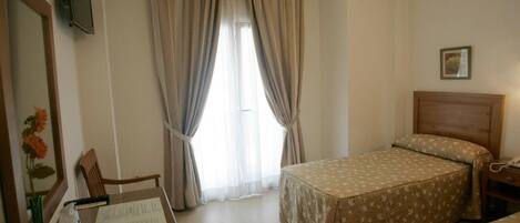 A hotel room with a bed, a desk with a chair, a window with curtains, and a framed picture on the wall.