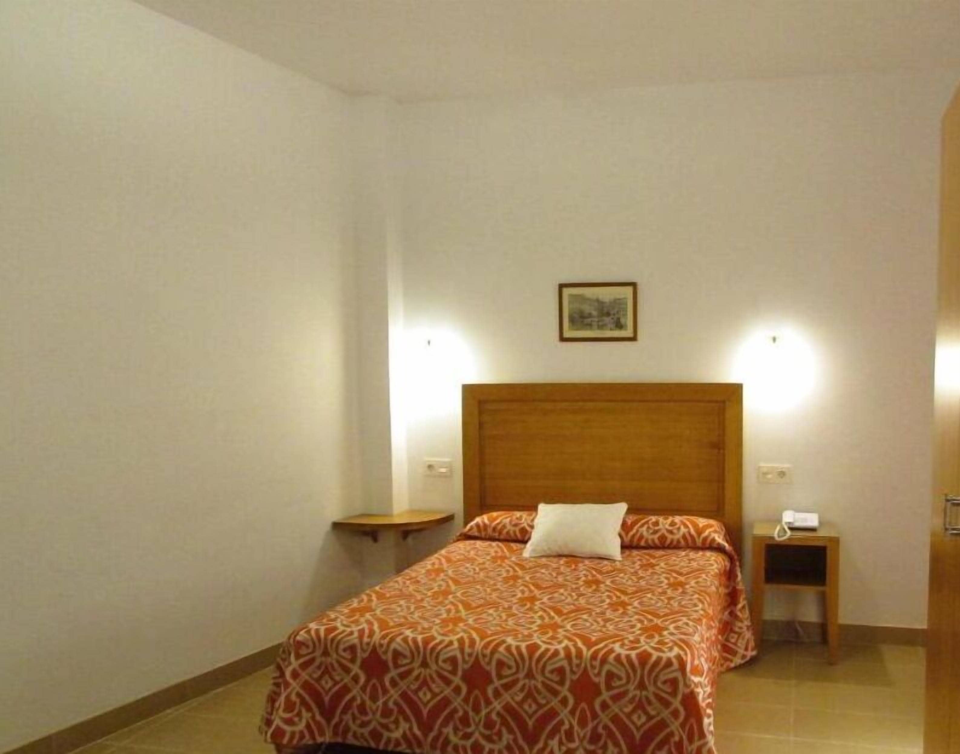 A neatly arranged bedroom with a wooden headboard, a single bed with patterned bedding, a bedside table with a lamp, and a small bedside table with a phone.