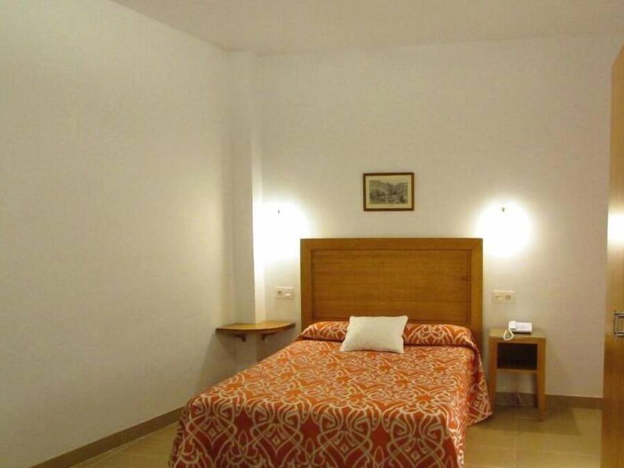 A neatly arranged bedroom with a wooden headboard, a single bed with patterned bedding, a bedside table with a lamp, and a small bedside table with a phone.