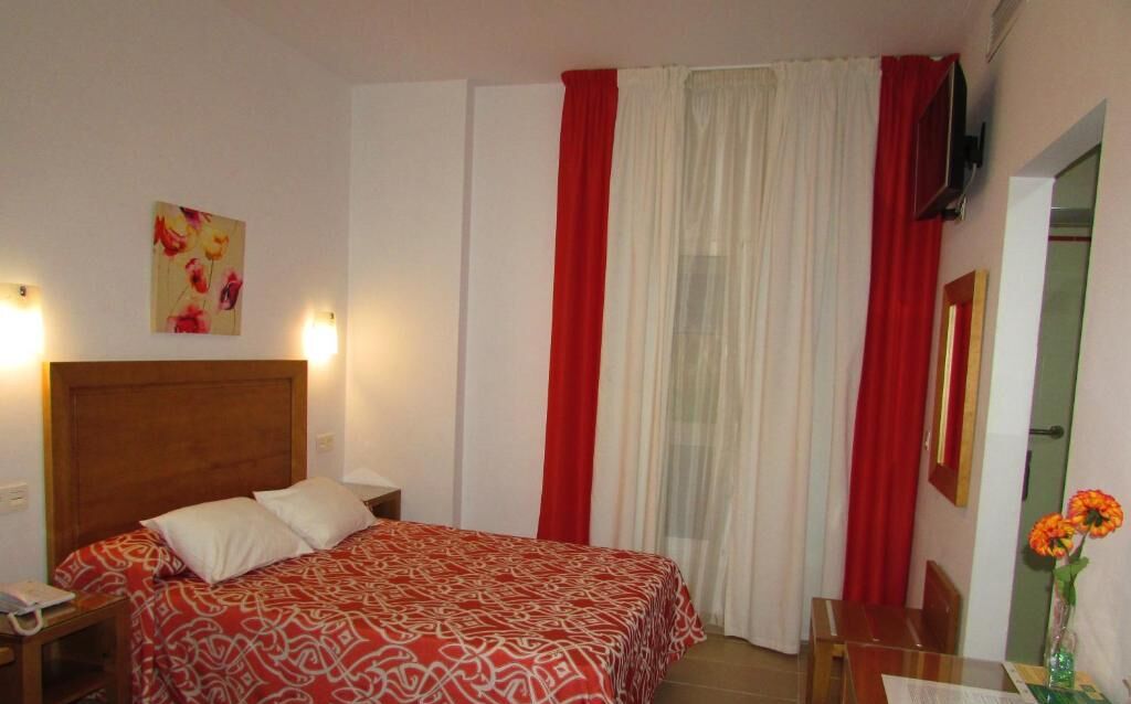 Quarto de hotel com uma cama, mesa de cabeceira, escrivaninha, cadeira, TV na parede e um quadro na parede.
