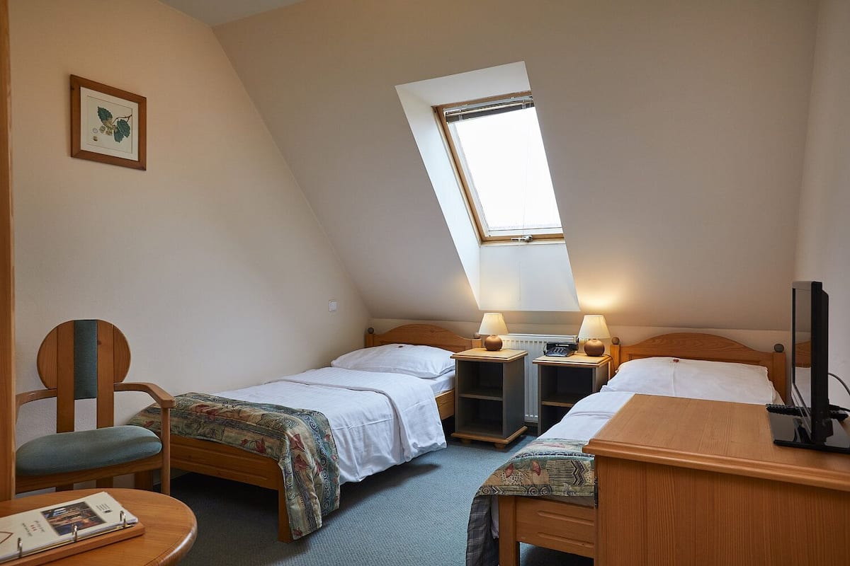 A small, well-lit room with two single beds, a wooden desk, a chair, and a small table. There is a window providing natural light and a framed picture on the wall.