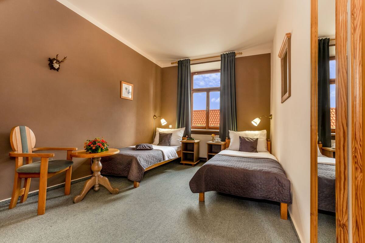 A hotel room with two single beds, a wooden chair, a small table with a flower arrangement, and a window with curtains.