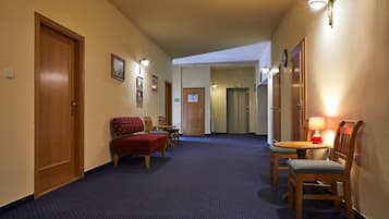 A hotel hallway with a blue carpet, wooden doors, a bench, chairs, and a table with a lamp.