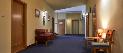 A hotel hallway with a blue carpet, wooden doors, a bench, chairs, and a table with a lamp.