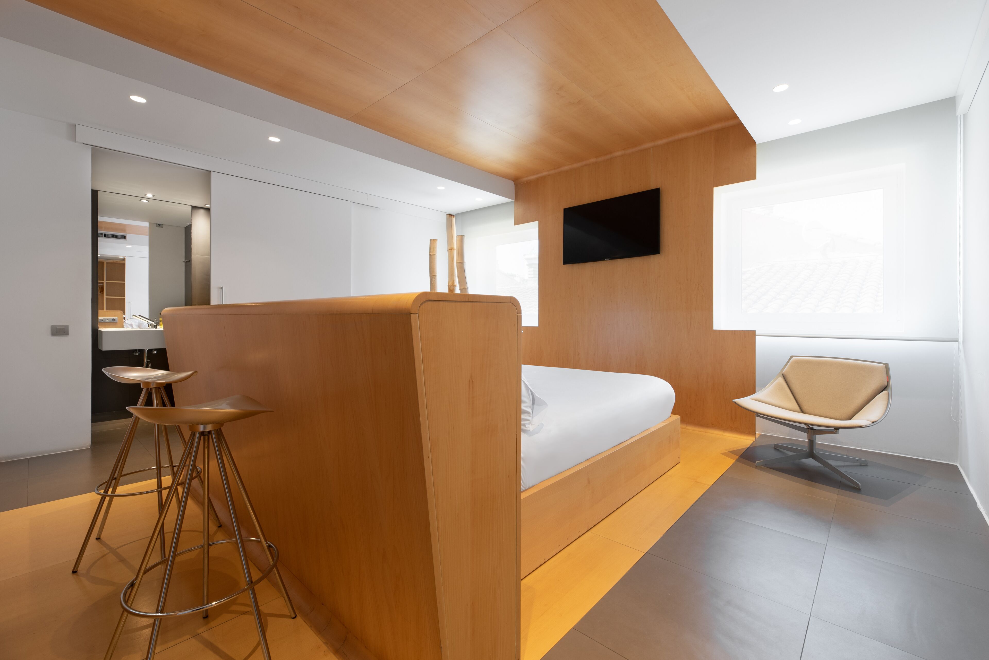 A modern hotel room with a wooden partition, a bed, a TV mounted on the wall, and a window providing natural light.