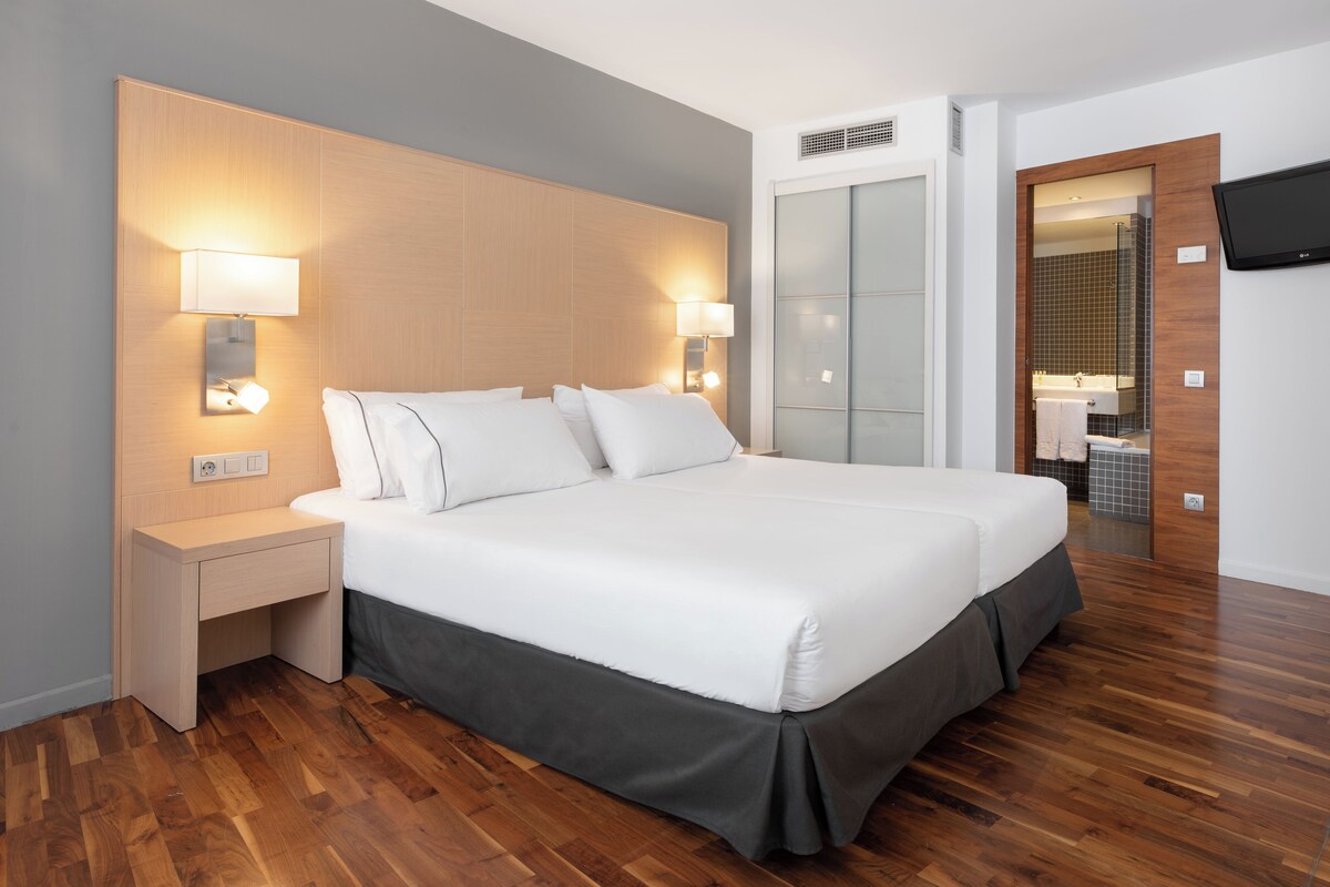 Une chambre d’hôtel moderne avec un grand lit, des lampes de chevet, une tête de lit en bois, un téléviseur fixé au mur et une salle de bain visible à travers une porte ouverte.