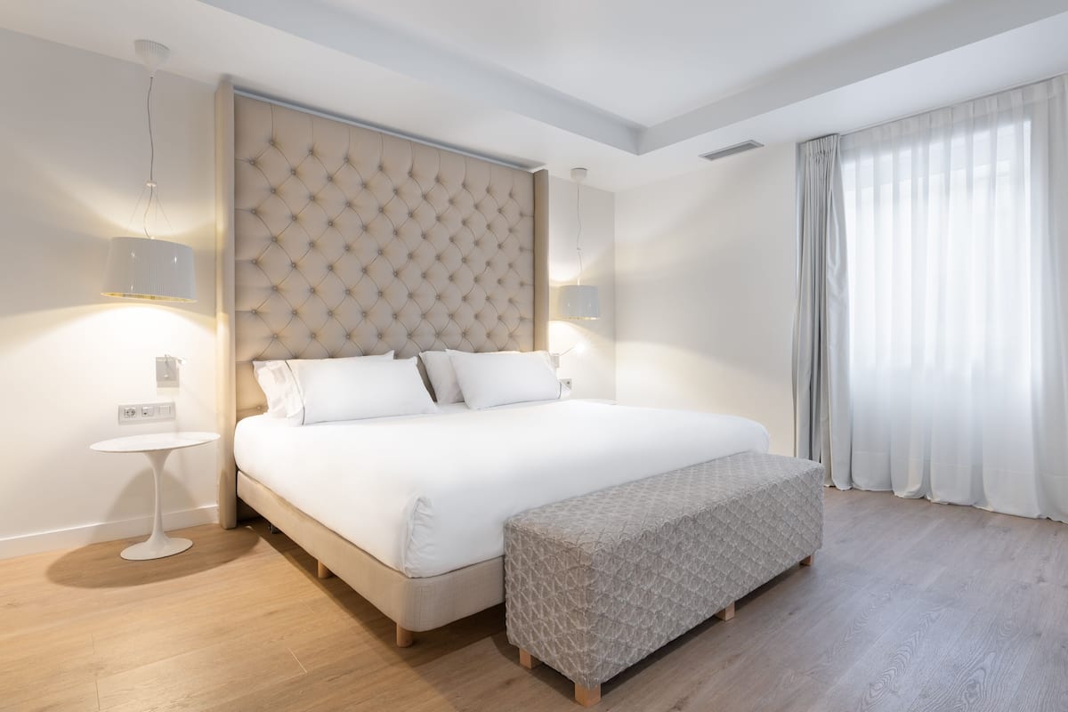 Een moderne hotelkamer met een groot bed, een nachtkastje, een bankje en een groot raam met gordijnen.
