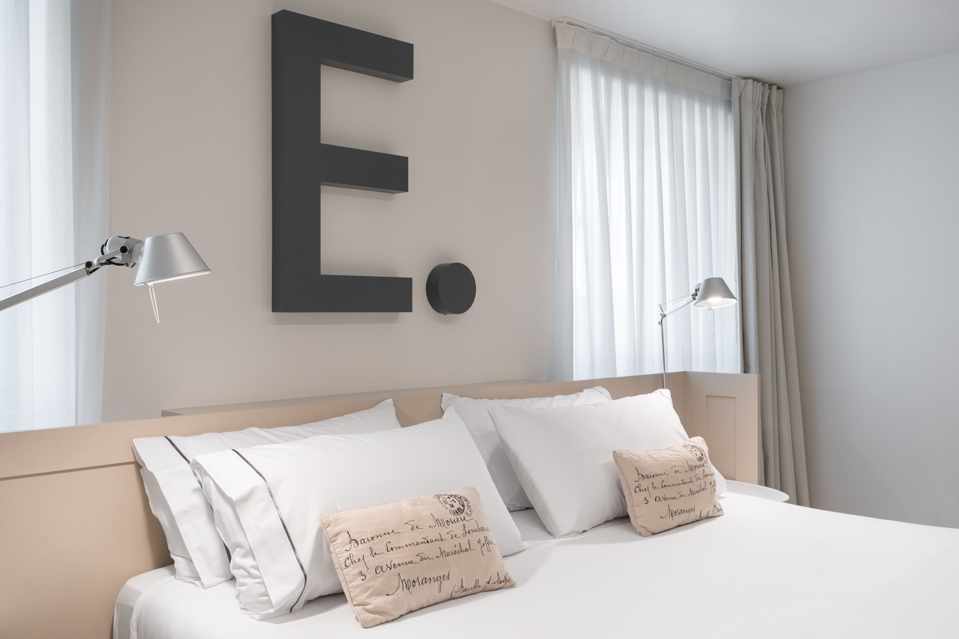 A neatly made bed with white linens and two decorative pillows, a large letter E on the wall, and two bedside lamps.