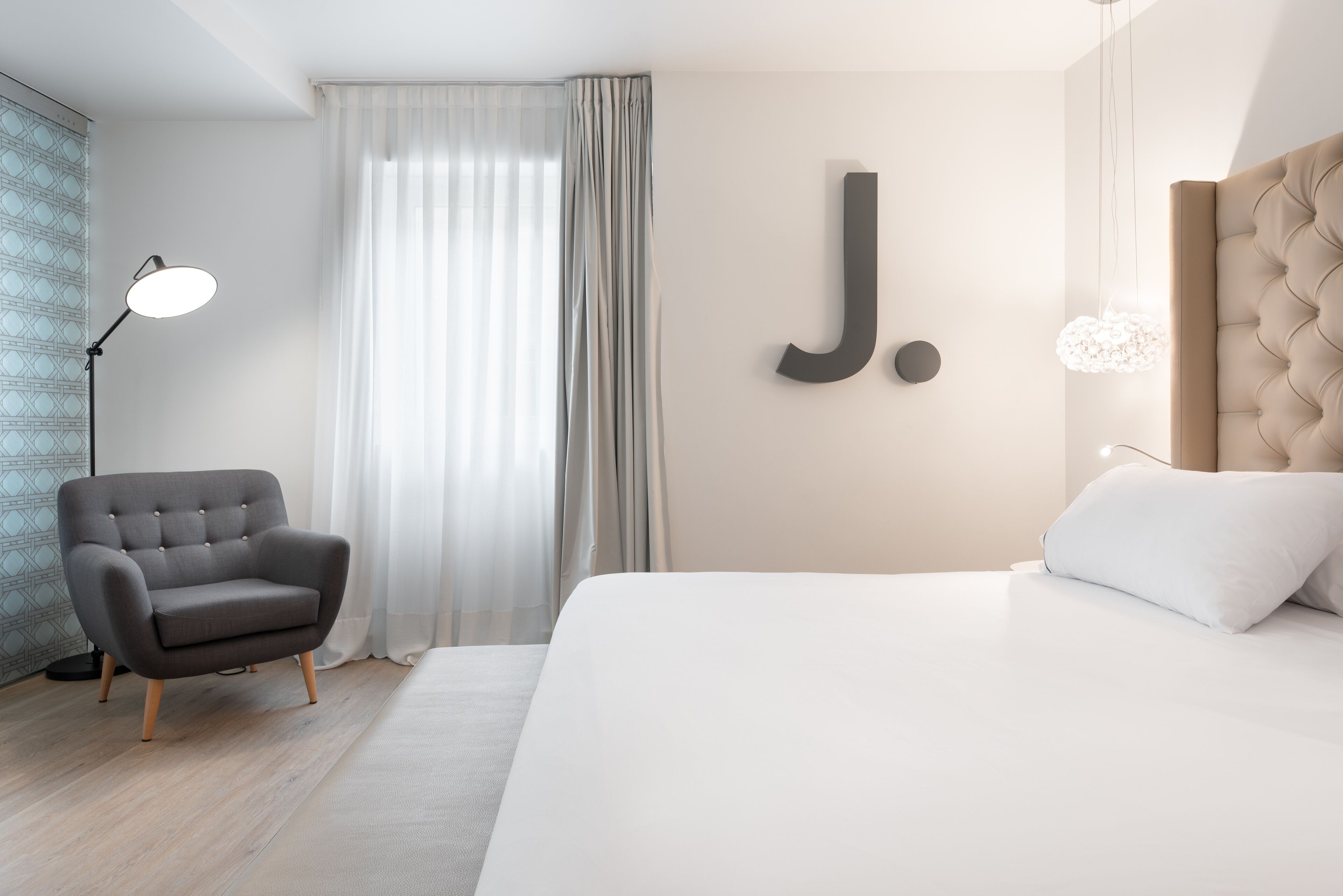 Una habitación de hotel moderna con una cama grande, un sillón gris, una lámpara de pie y una gran letra 'J' en la pared.