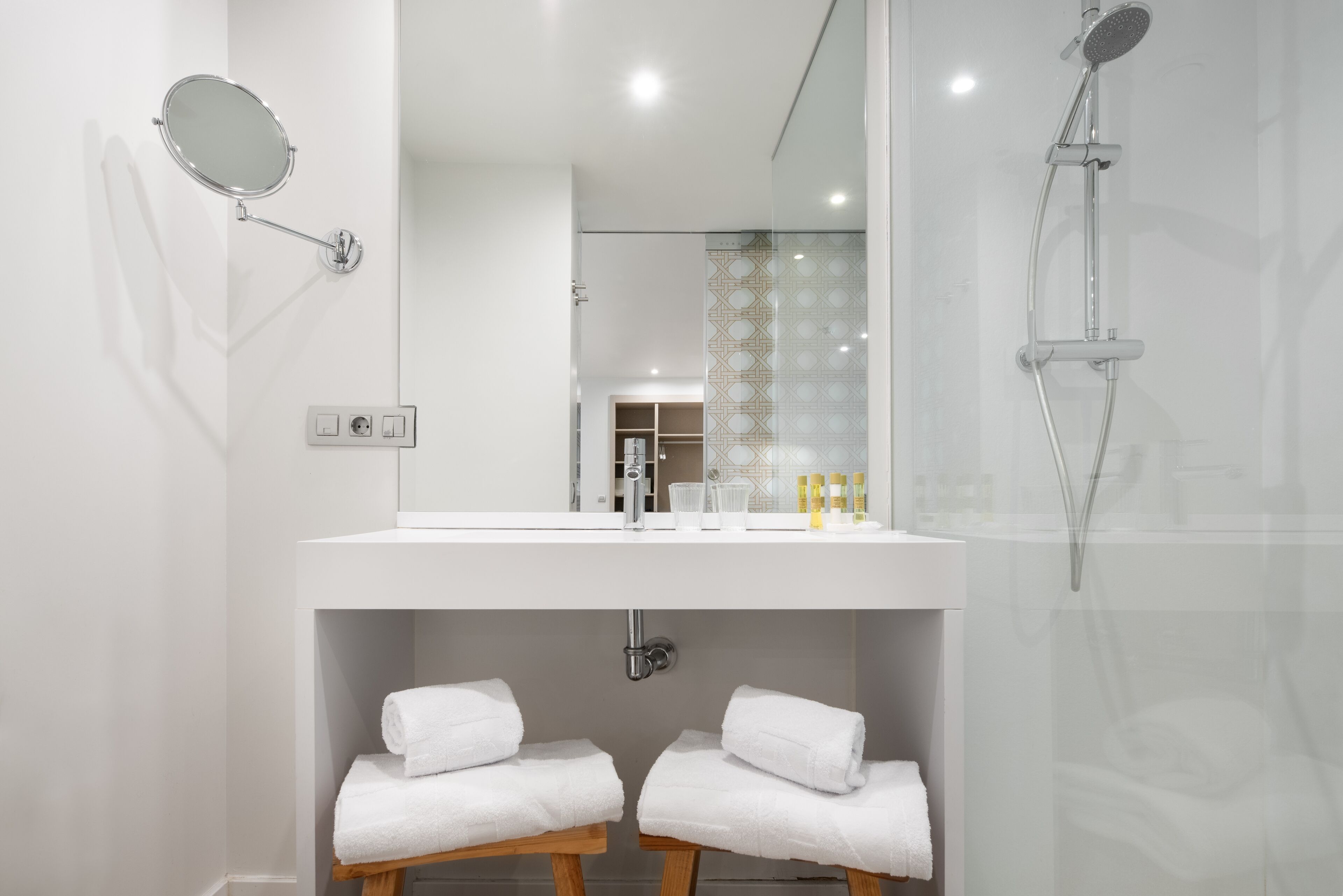 Un baño moderno con tocador blanco, un espejo grande, un cabezal de ducha de mano y dos taburetes de madera con toallas.