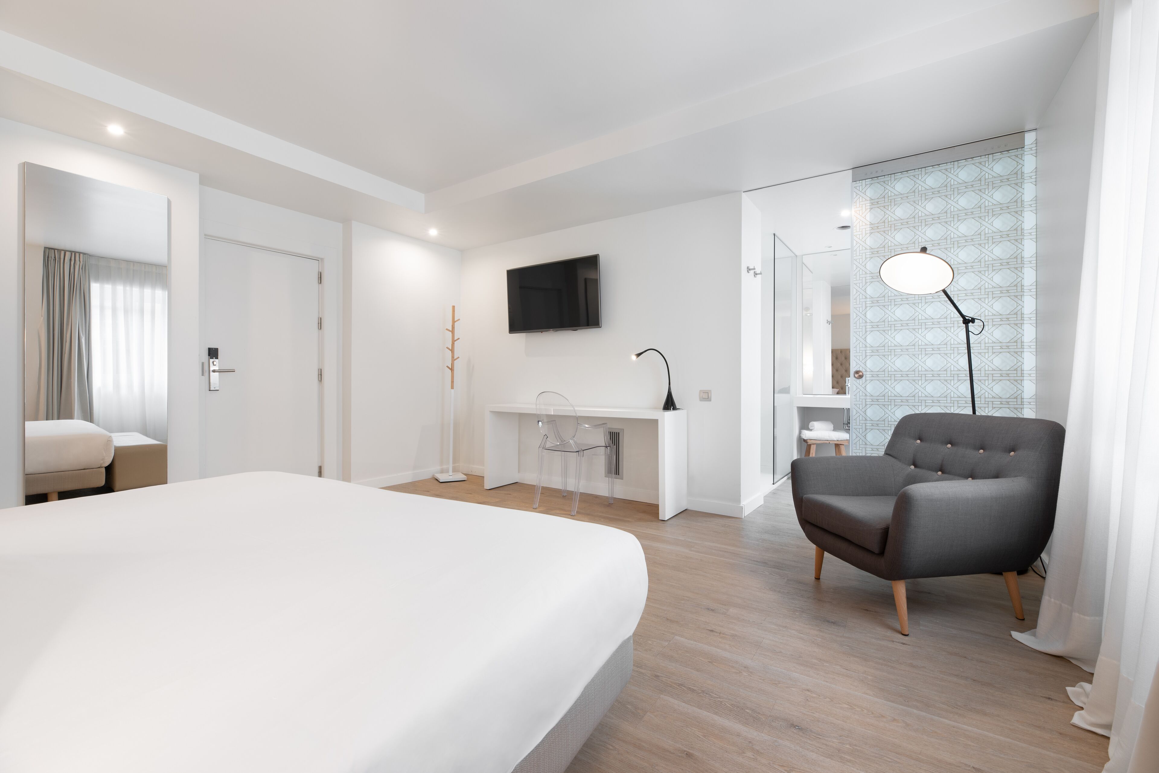 A modern hotel room with a large bed, a desk, a chair, a TV, and a lamp.