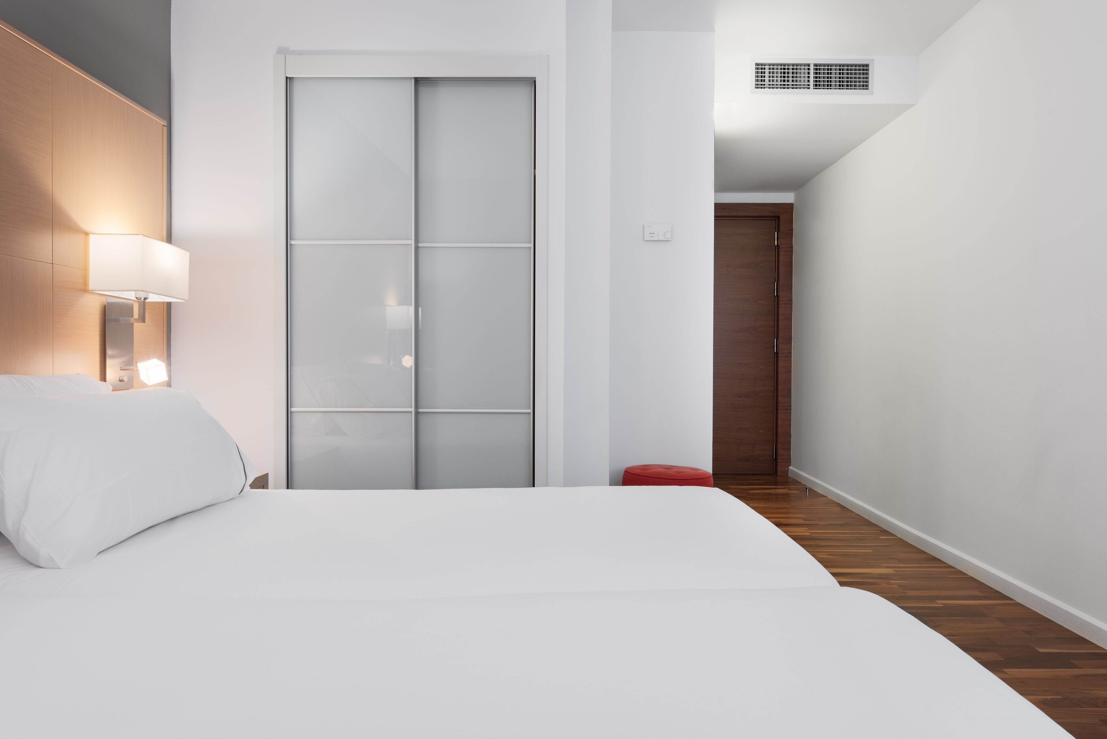 Habitación de hotel con cama, mesita de noche con lámpara, armario con puerta corredera de vidrio, reposapiés rojo y suelo de madera.