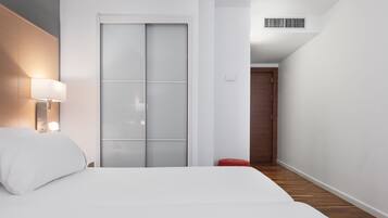 Habitación de hotel con cama, mesita de noche con lámpara, armario con puerta corredera de vidrio, reposapiés rojo y suelo de madera.