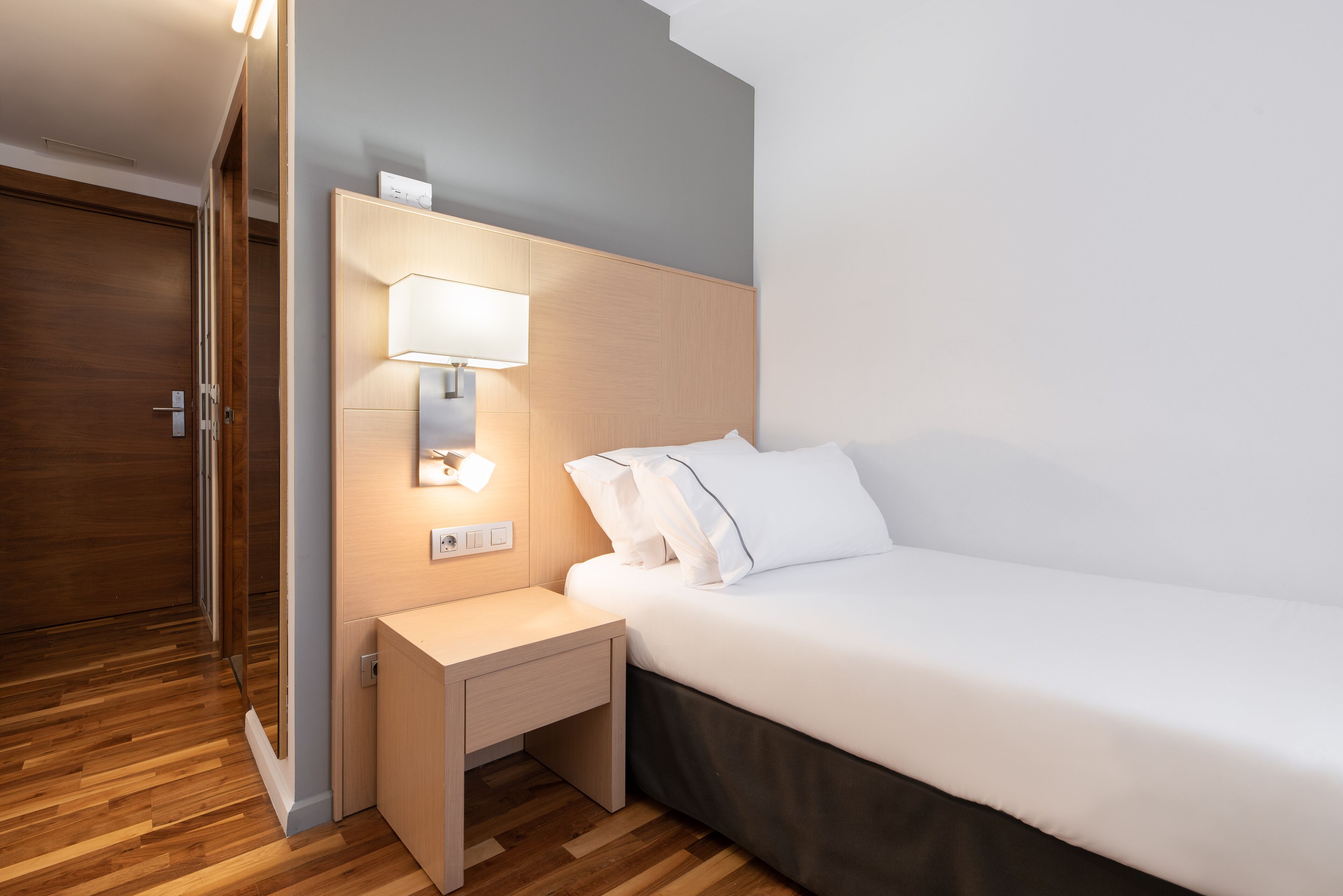 A modern hotel room with a wooden floor, a bed with white linens, a bedside table with a lamp, and a wooden wardrobe.