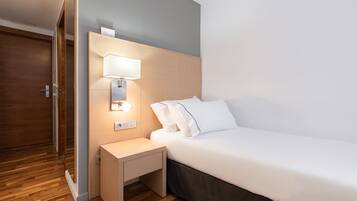 A modern hotel room with a wooden floor, a bed with white linens, a bedside table with a lamp, and a wooden wardrobe.