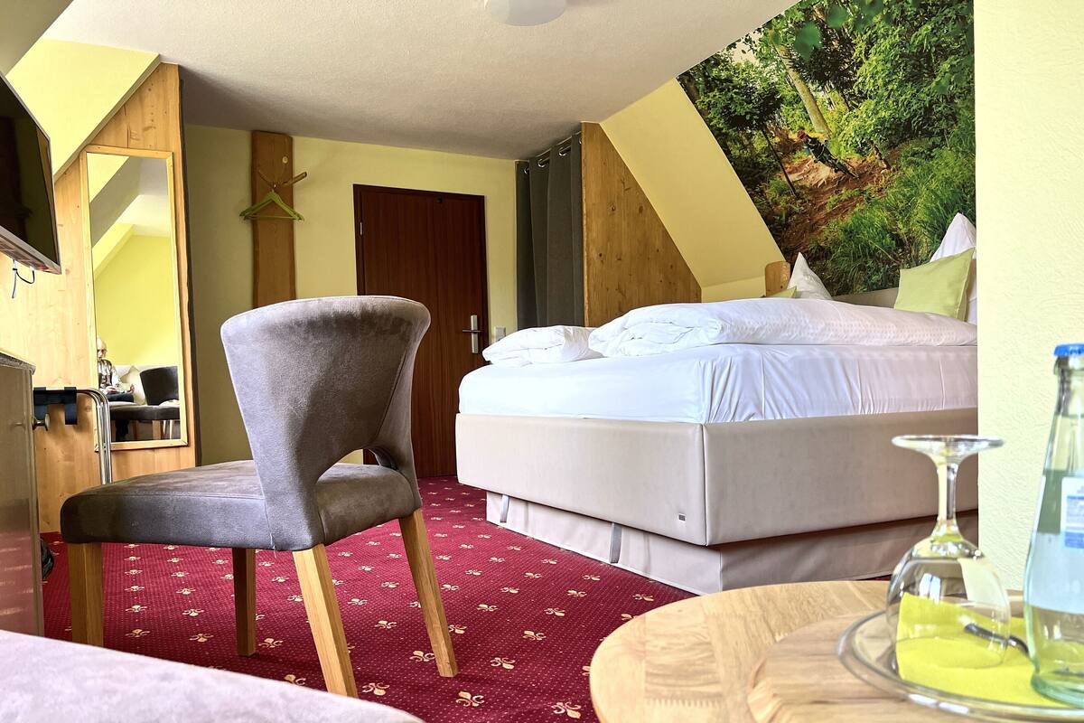 Una habitación de hotel con una cama, una silla, una mesa redonda con un vaso, y un cuadro de un bosque en la pared.