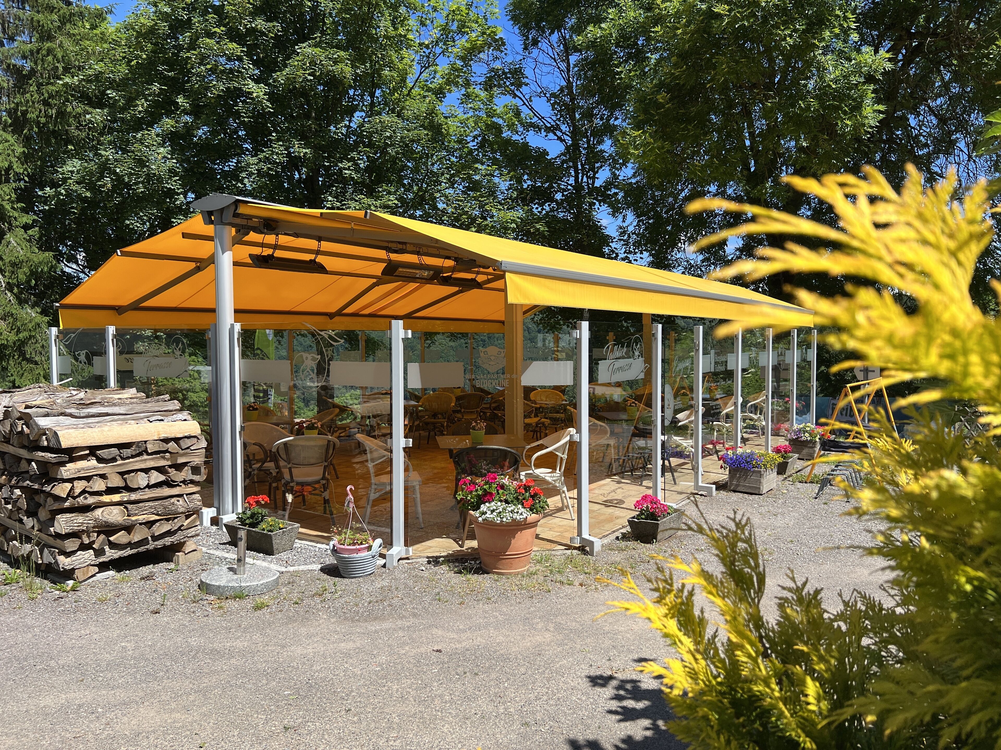 Un espace extérieur abrité, aménagé avec des tables et des chaises, entouré de plantes en pot et d’une pile de bois de chauffage.