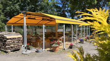 Un espace extérieur abrité, aménagé avec des tables et des chaises, entouré de plantes en pot et d’une pile de bois de chauffage.