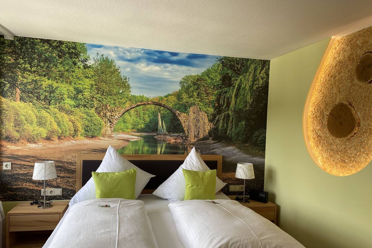 Un dormitorio con un mural de un bosque y un puente, dos camas con almohadas blancas y verdes, lámparas de noche y una mesita de noche de madera con una radio.