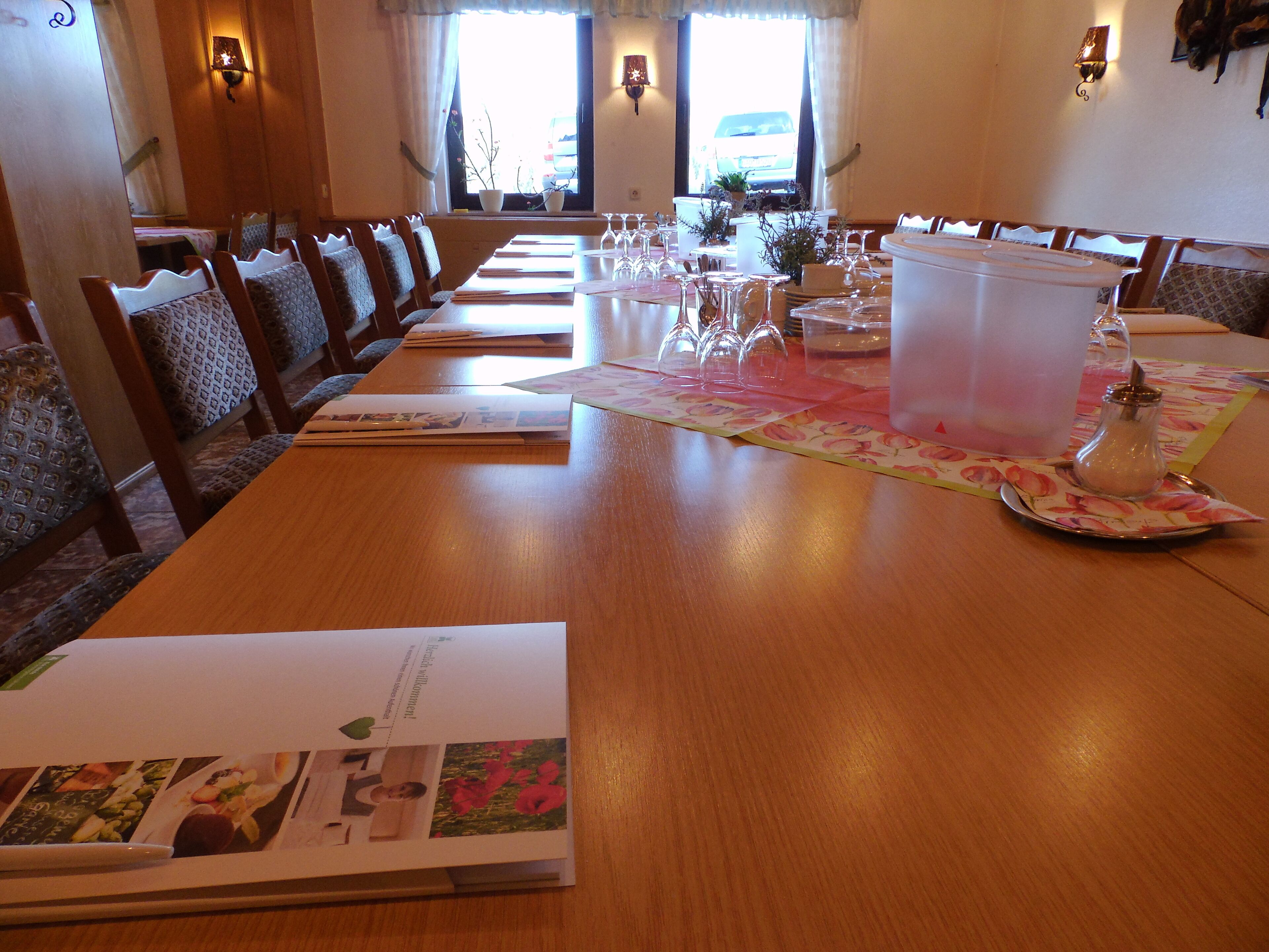A dining area with a long wooden table set for a meal, chairs with patterned cushions, and a view of the outside through large windows.