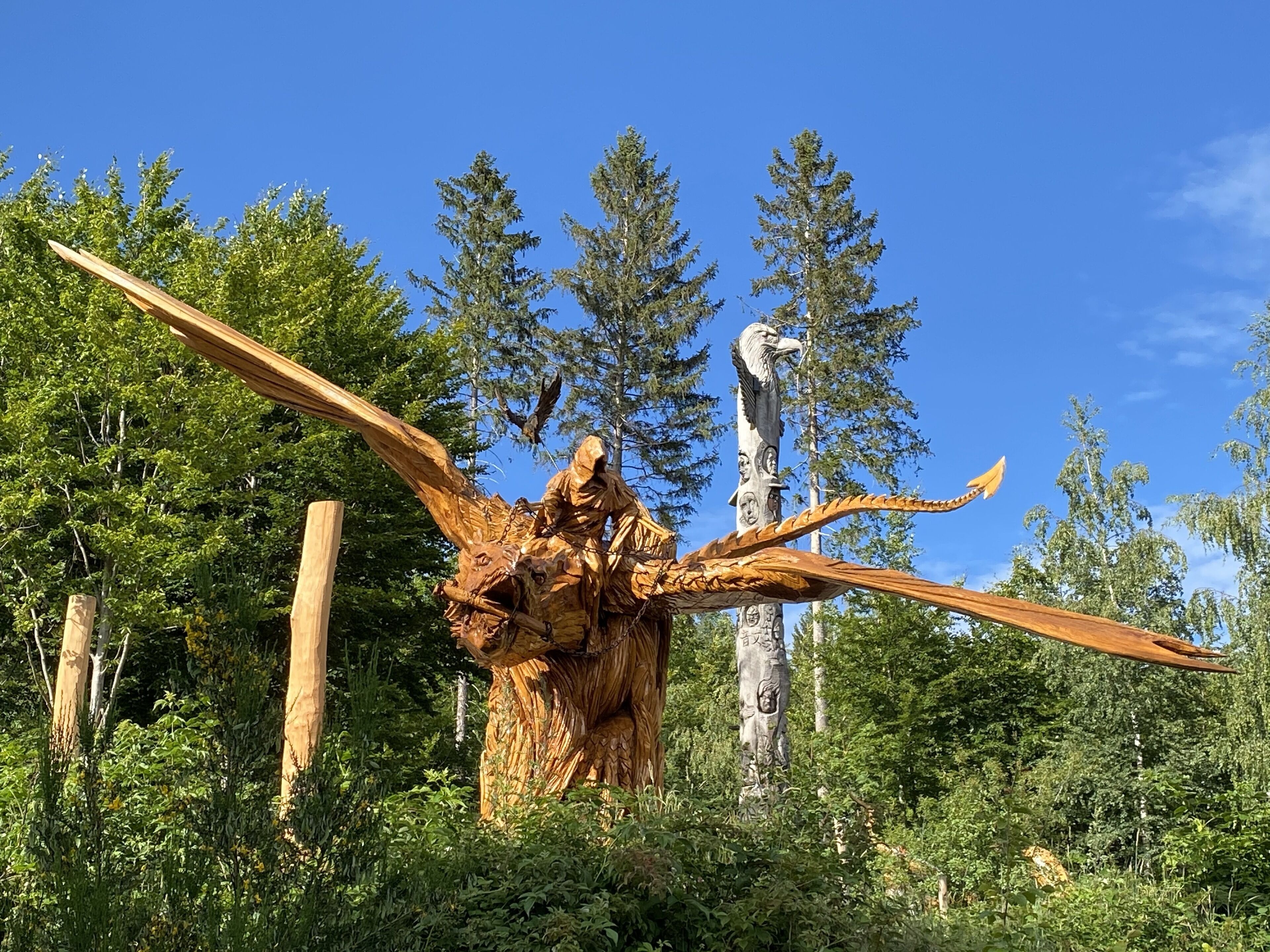 Eine große Holzskulptur einer Figur mit ausgestreckten Armen, umgeben von dichtem Grün und hohen Bäumen.