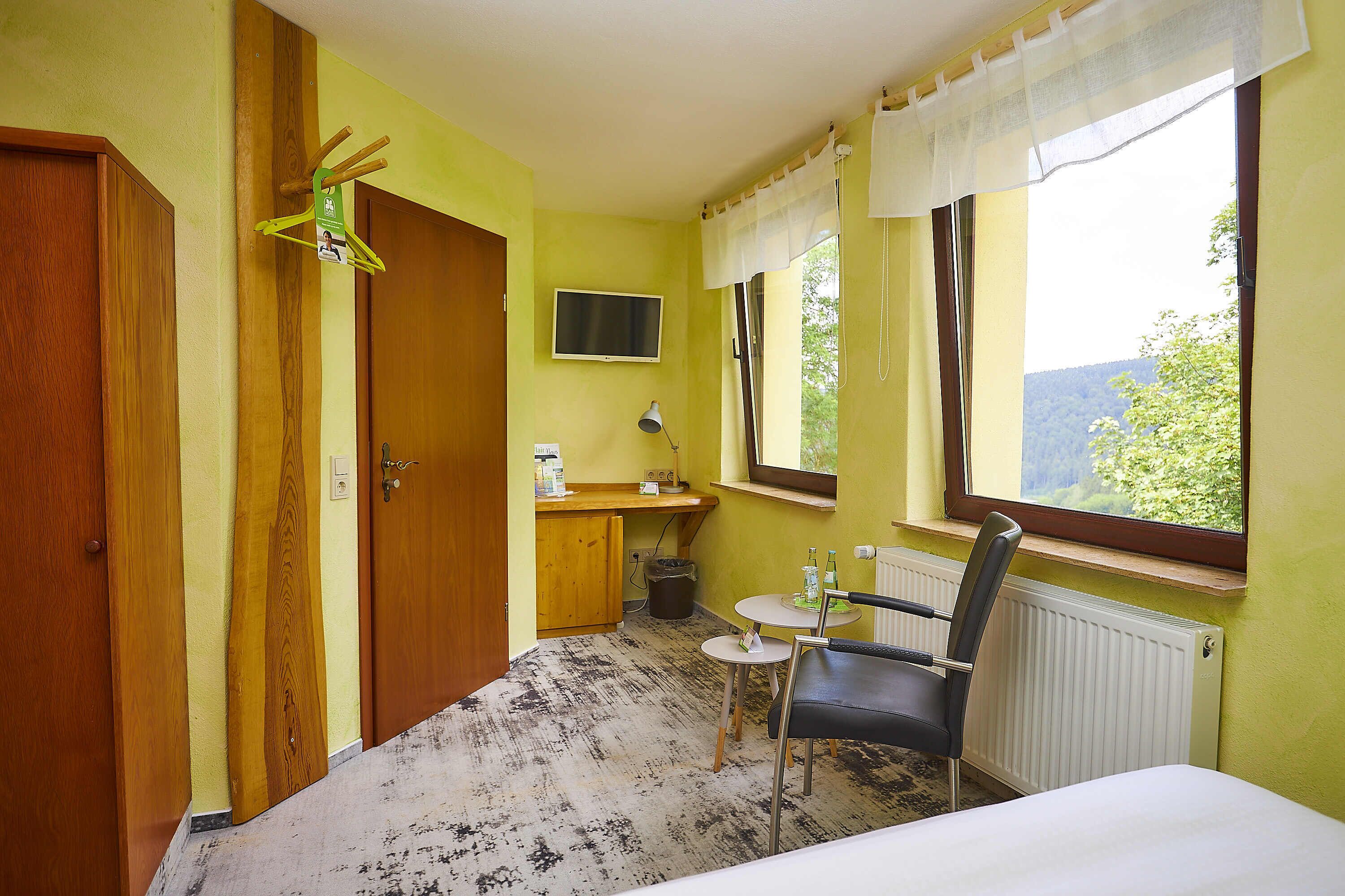 Ein kleines Zimmer mit Bett, Stuhl, Tisch, Fernseher, Fenster und Tür.