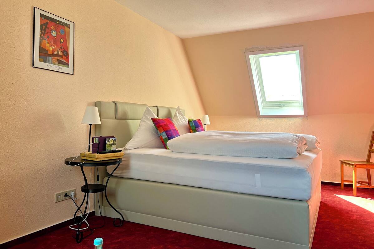 Una habitación de hotel con cama, un escritorio con lámpara, una silla, una ventana y un cuadro en la pared.