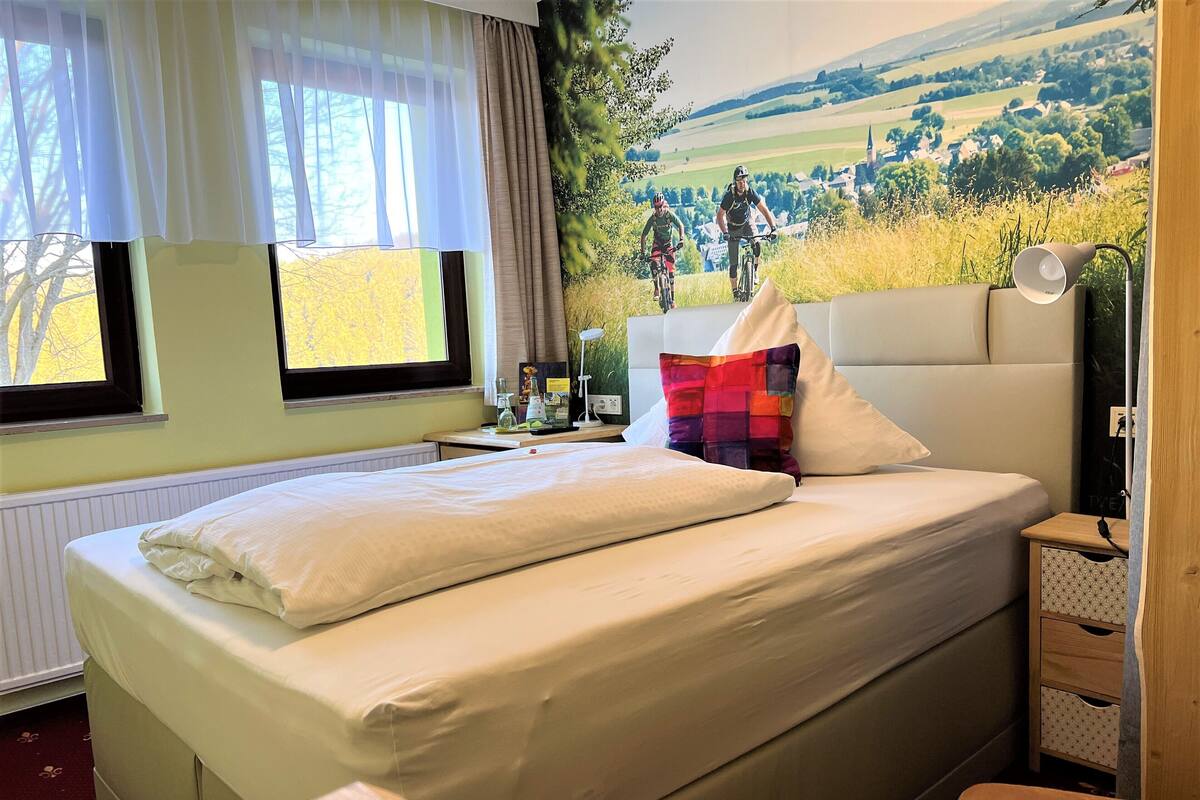 Habitación de hotel con cama, mesita de noche, lámpara y un mural con ciclistas en un paisaje pintoresco.